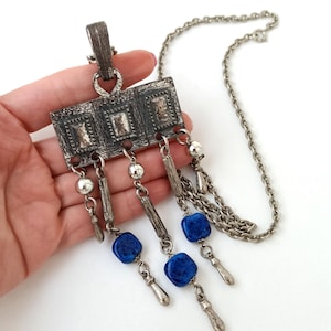 May include: A silver tone necklace with a rectangular pendant featuring a geometric design and blue stone accents.