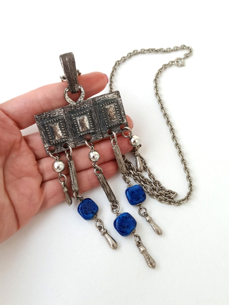 May include: A silver tone necklace with a rectangular pendant featuring a geometric design and blue stone accents.