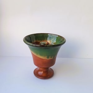 Peut inclure: Un vase en céramique fait à la main avec un dessus évasé et une base en piédestal. Le vase présente un dégradé de couleurs, passant d'un bord vert foncé à une bande orange et une base marron. Un petit objet rond repose à l'intérieur.