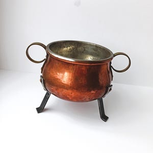 May include: A small, antique copper cauldron with a silver-colored interior. It has two brass handles and rests on three black metal legs. The cauldron's exterior has a rich, reddish-brown patina.