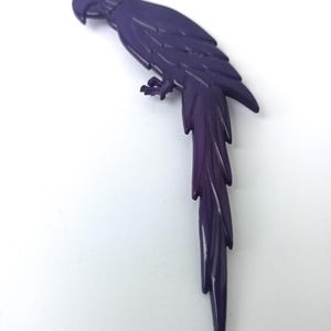 May include: A purple plastic parrot brooch with a rhinestone eye. The parrot is perched with its tail feathers fanned out.