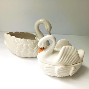 Puede incluir: Dos cuencos de cerámica blanca con forma de cisne. El cisne más grande es un cuenco abierto con detalles de alas texturizadas. El cisne más pequeño cuenta con una tapa extraíble con un diseño de ala esculpida y un pico pintado de naranja.
