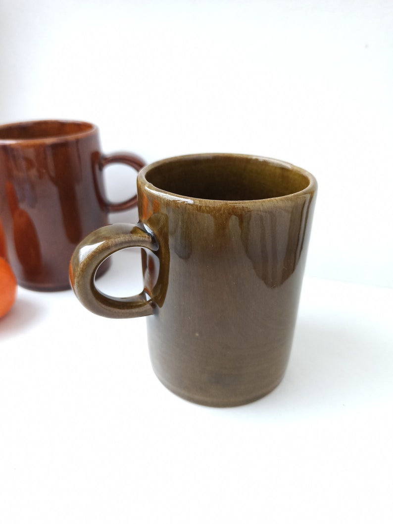 Puede incluir: Dos tazas de cer&aacute;mica, una marr&oacute;n y otra verde oscuro, con asas. Las tazas est&aacute;n sobre una superficie blanca.