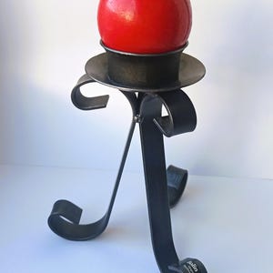 Könnte beinhalten: Ein dekorativer Kerzenhalter aus schwarzem Metall mit einer runden roten Kerze. Der Kerzenhalter hat einen kreisförmigen Sockel und ein geschwungenes, scrollartiges Design. Die Kerze hat einen Durchmesser von ca. 7.6 cm.