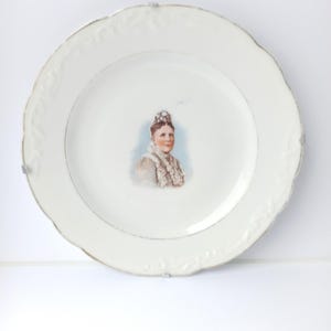 May include: White porcelain plate with a gold rim and a raised floral design. The center of the plate features a printed image of a woman wearing a crown and a white dress.