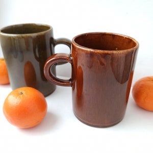 Puede incluir: Dos tazas de cer&aacute;mica, una marr&oacute;n y otra verde, con un asa marr&oacute;n. Las tazas est&aacute;n sobre una superficie blanca con tres peque&ntilde;as naranjas delante de ellas.
