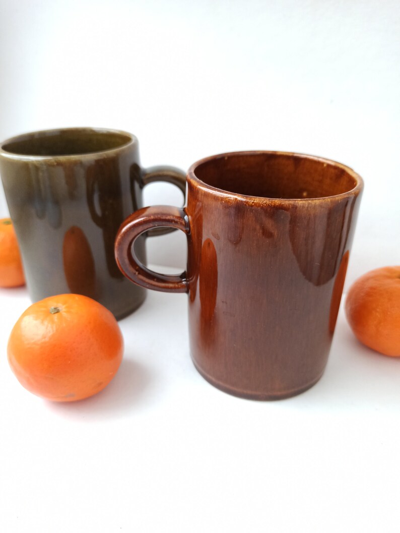Puede incluir: Dos tazas de cer&aacute;mica, una marr&oacute;n y otra verde, con un asa marr&oacute;n. Las tazas est&aacute;n sobre una superficie blanca con tres peque&ntilde;as naranjas delante de ellas.