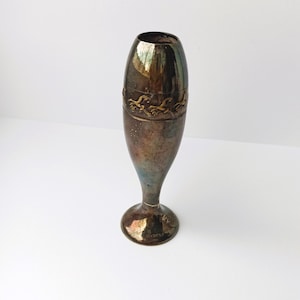 May include: A vintage silver-toned vase with a flared base and a narrow neck. The vase has a decorative band with a repeating bird or wave pattern. The metal has a tarnished appearance, giving it a rustic charm.