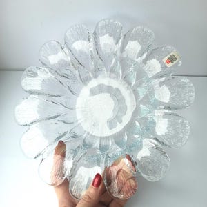 May include: Clear glass flower-shaped bowl with textured petals. The bowl has a central, circular design and is held by a hand. The glass has a slightly blue tint. A small tag is visible on one of the petals.