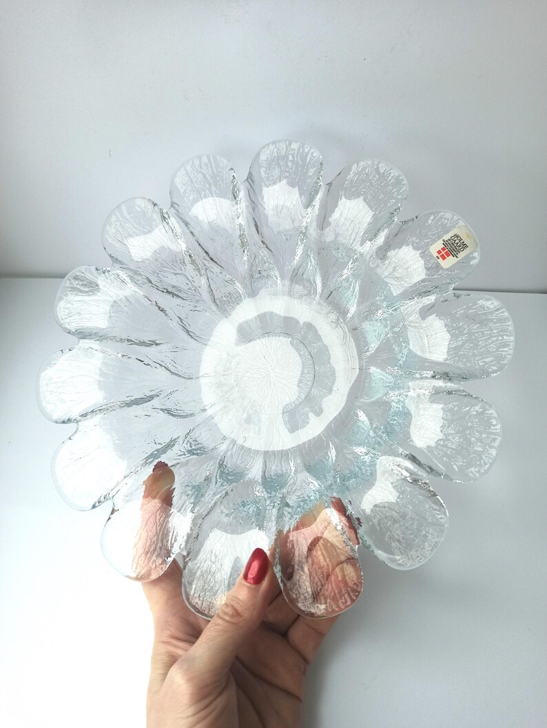 May include: Clear glass flower-shaped bowl with textured petals. The bowl has a central, circular design and is held by a hand. The glass has a slightly blue tint. A small tag is visible on one of the petals.