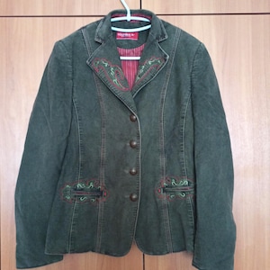 May include: A dark green blazer with decorative red and green embroidery around the collar and pockets. The blazer has a button-up front and a red striped lining. The blazer is hanging on a white hanger.