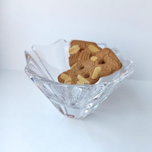 Peut inclure: Un bol en verre transparent en forme d'étoile contient deux biscuits aux amandes. Les biscuits sont brun clair avec des tranches d'amandes. Le bol a un design texturé et repose sur une surface blanche.