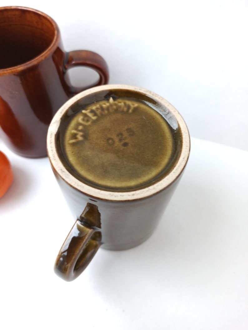 Puede incluir: Una taza de cer&aacute;mica marr&oacute;n con asa. La base de la taza tiene un esmalte verde con las palabras "W. Germany" y el n&uacute;mero "026" impresos en ella.