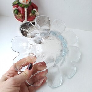 May include: Clear glass flower-shaped bowl with a textured surface. The bowl is held by a hand with dark nail polish. A snowman figurine in a red coat and hat is in the background.