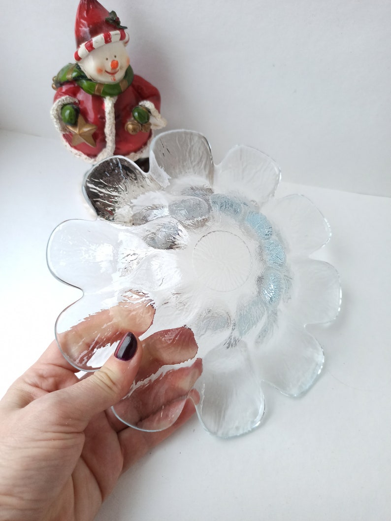 May include: Clear glass flower-shaped bowl with a textured surface. The bowl is held by a hand with dark nail polish. A snowman figurine in a red coat and hat is in the background.