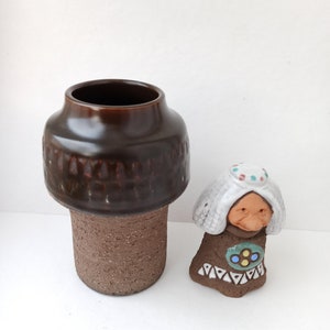 以下が含まれることがあります： 茶色のテクスチャ加工された表面を持つセラミックの花瓶と、白い髪と茶色のドレスを着た女性の小さなセラミックの置物。女性は青と緑のディテールで描かれた顔をしています。