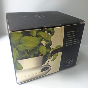 Peut inclure: Une boîte noire avec une image d'un pot à herbes blanc avec du basilic vert et de l'eau qui coule d'un bec. La boîte porte l'inscription "HOLD herb pot" et d'autres traductions.
