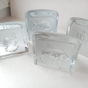 May include: Four clear glass square coasters with etched zodiac symbols: a scorpion, a fish, a bull, and a woman. The coasters are made of thick glass and have a frosted appearance where the zodiac symbols are etched.
