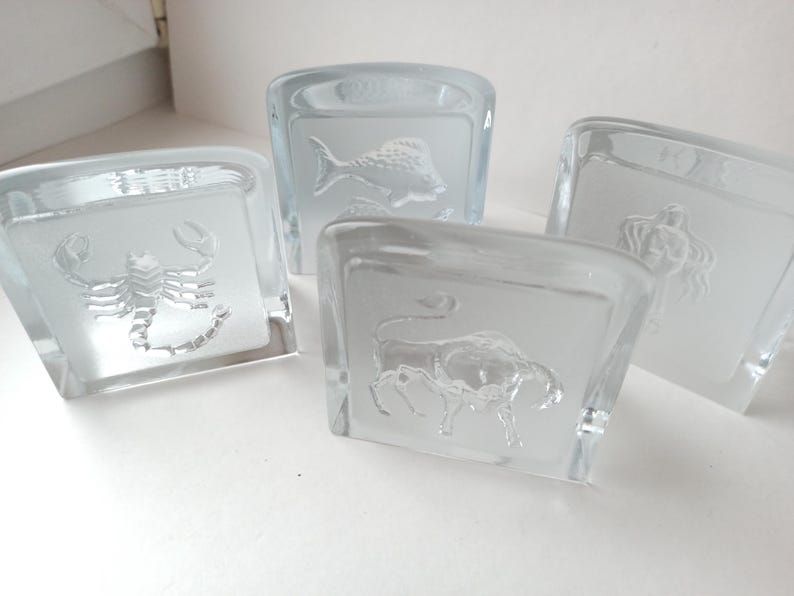 May include: Four clear glass square coasters with etched zodiac symbols: a scorpion, a fish, a bull, and a woman. The coasters are made of thick glass and have a frosted appearance where the zodiac symbols are etched.