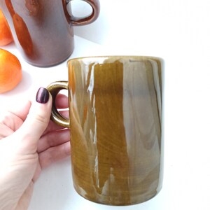 Puede incluir: Dos tazas de cer&aacute;mica marr&oacute;n con un acabado brillante. Una taza se sostiene en una mano, mientras que la otra est&aacute; sobre una superficie blanca.