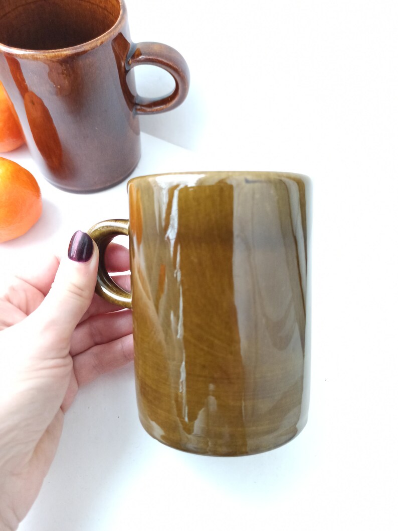 Puede incluir: Dos tazas de cer&aacute;mica marr&oacute;n con un acabado brillante. Una taza se sostiene en una mano, mientras que la otra est&aacute; sobre una superficie blanca.