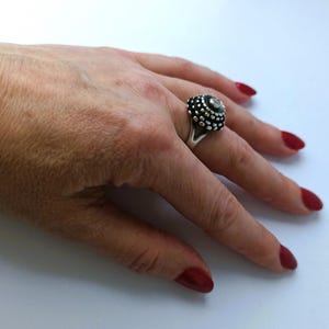 May include: A silver ring with a domed, textured design featuring small, round accents. The ring is worn on a hand with red nail polish.