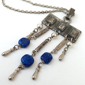May include: A silver necklace with a rectangular pendant featuring a geometric design. The pendant has three blue gemstone beads hanging from it.