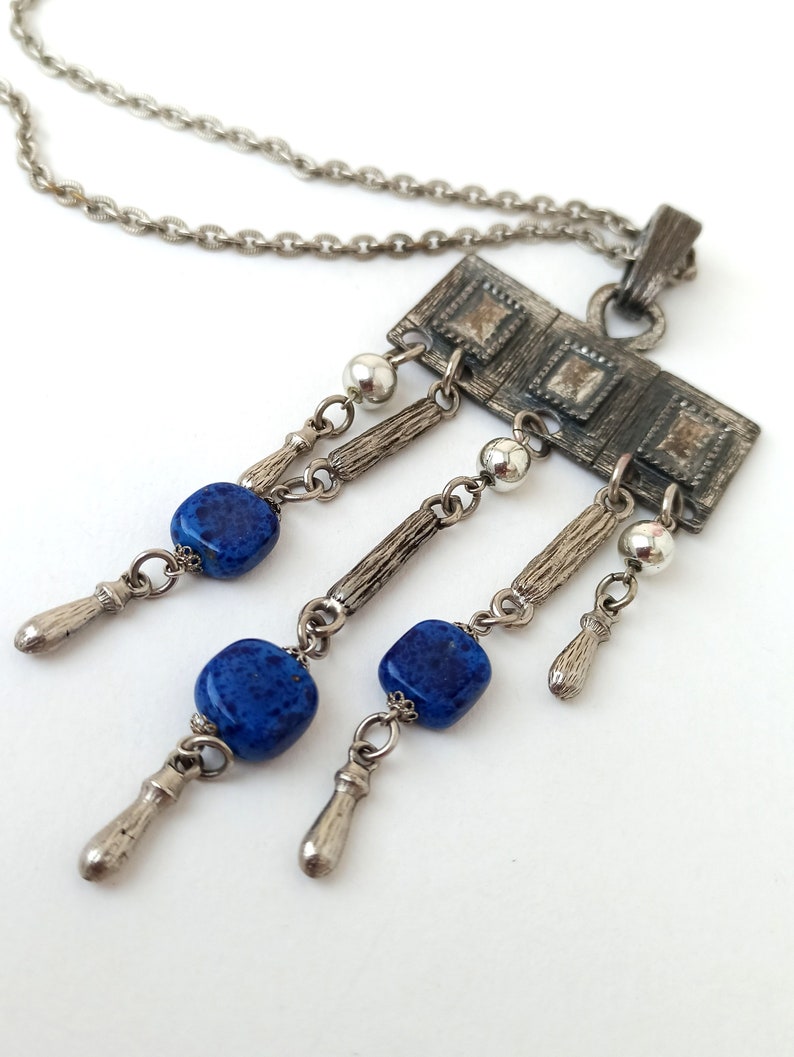 May include: A silver necklace with a rectangular pendant featuring a geometric design. The pendant has three blue gemstone beads hanging from it.