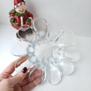 May include: A clear glass flower-shaped bowl with petal-like edges. The bowl is held by a hand with dark red nail polish. A snowman figurine in a red coat and hat is in the background.