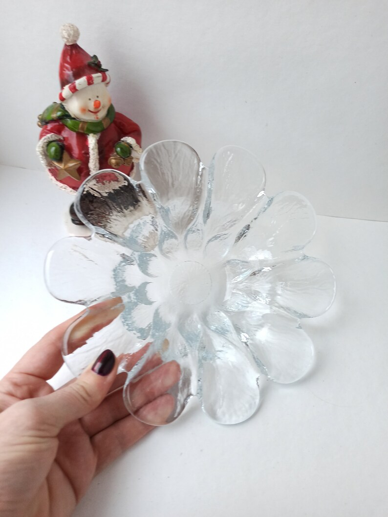 May include: A clear glass flower-shaped bowl with petal-like edges. The bowl is held by a hand with dark red nail polish. A snowman figurine in a red coat and hat is in the background.