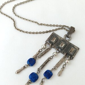 May include: A silver chain necklace with a rectangular pendant featuring a geometric design and blue stone accents.