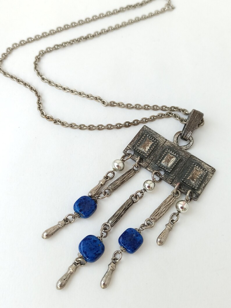 May include: A silver chain necklace with a rectangular pendant featuring a geometric design and blue stone accents.