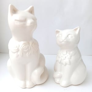 May include: Two white ceramic cat figurines. The larger cat is sitting with its tail curled around its legs. The smaller cat is sitting with its tail straight out behind it. Both cats have a floral design around their necks.