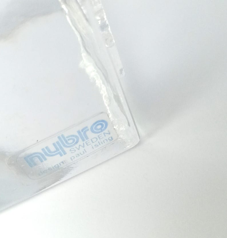 May include: A clear glass object with a label that reads "nybro SWEDEN design: paul isling". The object has a slightly textured surface and is partially visible against a white background.