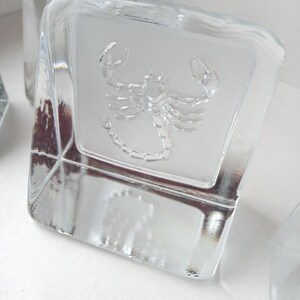 May include: Clear glass cube with a frosted panel featuring an embossed scorpion design. The cube has a square shape and is made of transparent glass. The scorpion is detailed and centered on the panel.