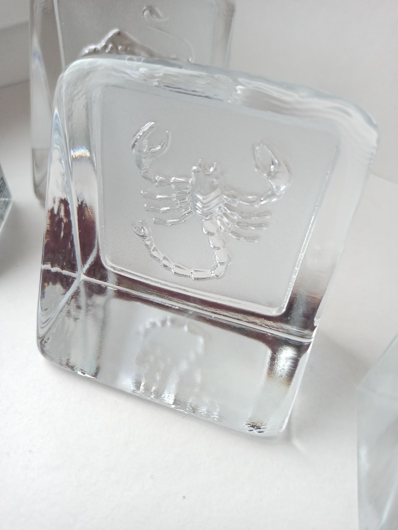 May include: Clear glass cube with a frosted panel featuring an embossed scorpion design. The cube has a square shape and is made of transparent glass. The scorpion is detailed and centered on the panel.
