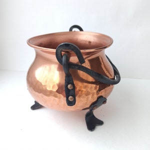 Könnte beinhalten: Ein kleiner Kupfertopf mit gehämmerter Oberfläche und schwarzen Eisenhenkeln und -beinen.