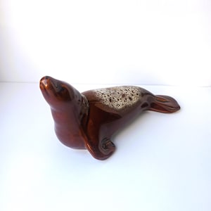 Könnte beinhalten: Eine braune Keramik-Seehundfigur mit glänzendem Finish. Der Seehund liegt mit erhobenem Kopf. Die Oberseite des Seehunds hat ein strukturiertes, helles Muster.