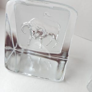 May include: Clear glass square-shaped object with a frosted panel featuring a detailed embossed bull. The bull is depicted with horns and a raised tail. The object is likely a decorative piece or a paperweight.