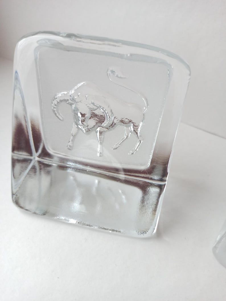May include: Clear glass square-shaped object with a frosted panel featuring a detailed embossed bull. The bull is depicted with horns and a raised tail. The object is likely a decorative piece or a paperweight.