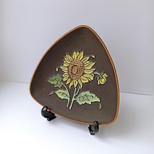 Può includere: Un piatto triangolare in ceramica marrone con un motivo a girasole. Il girasole è giallo e marrone, con foglie verdi e uno stelo marrone. Il piatto ha un bordo marrone chiaro ed è esposto su un supporto nero.