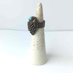 May include: A silver ring with a turquoise stone set in a detailed, ornate design. The ring is displayed on a white cone-shaped ring holder.