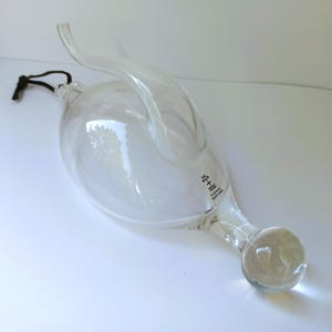 May include: A clear, hand-blown glass vessel with a curved, horn-like spout and a spherical base. A black cord is attached to the top. The glass features a white swirl pattern and a small printed label that reads "Skruf Sweden Exclusively hand crafted."