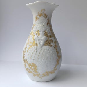 May include: White ceramic vase with a ruffled top, featuring a raised relief design of a woman in a gown, accented with gold detailing. The vase has a classic shape and is suitable for floral arrangements or as a decorative piece.