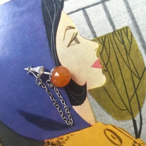 Könnte beinhalten: Ein einzelner Ohrring mit einem runden, orangefarbenen Edelstein und einer Silberkette. Der Ohrring ist vor einer Vintage-Illustration eines Frauenprofils platziert, mit einem Blau- und Gelbschema.