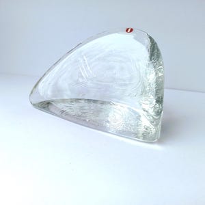 Può includere: Scultura in vetro trasparente dalla forma triangolare arrotondata. Il vetro presenta motivi a spirale incisi e un piccolo punto rosso visibile sulla parte superiore. La scultura è esposta su una superficie bianca.