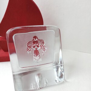 May include: A clear, rectangular glass block with a frosted panel featuring a stylized red figure. The figure has flowing hair and a detailed torso. A red, curved object is partially visible in the background.