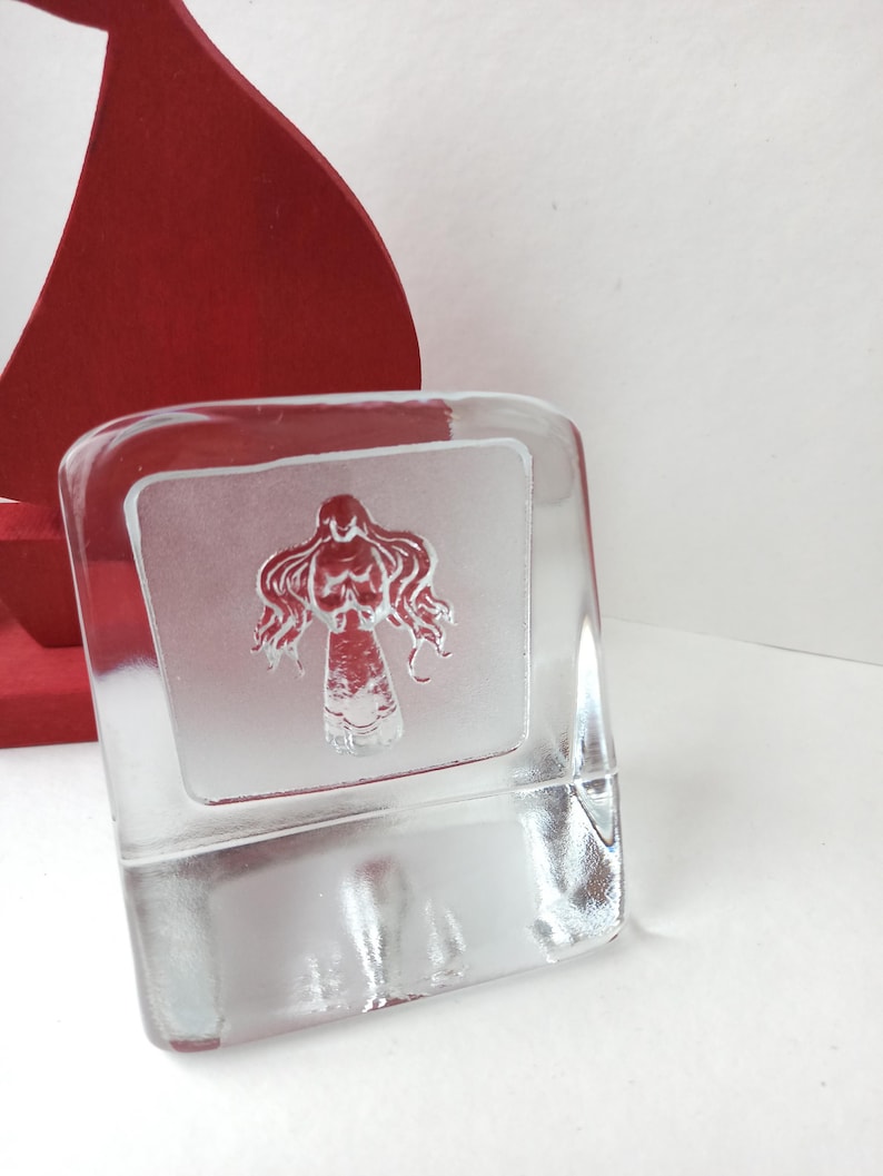 May include: A clear, rectangular glass block with a frosted panel featuring a stylized red figure. The figure has flowing hair and a detailed torso. A red, curved object is partially visible in the background.