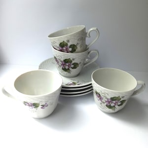 May include: A set of white porcelain teacups and saucers. The cups and saucers are decorated with a floral pattern of purple violets and green leaves. The teacups have a textured surface and curved handles. The set includes four cups and saucers.