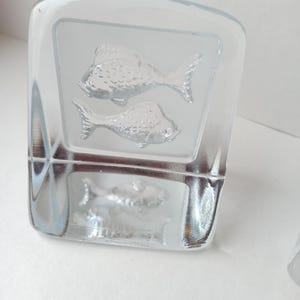 May include: A clear glass block with etched fish designs. Two fish are etched on the front, and one on the side. The glass has a frosted appearance where the fish are etched, creating a contrast with the clear glass.